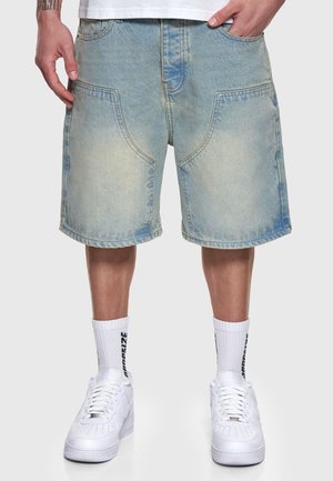 Mann trägt hellblaue Jeansshorts, weiße Sneakers, weiße Sportsocken mit schwarzem Text, steht mit den Händen nahe den Taschen vor einem einfarbigen Hintergrund.