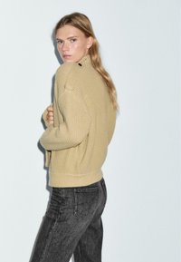 Maglione a coste beige con collo alto e dettagli con bottoni laterali, abbinato a jeans neri. Il tessuto appare morbido e testurizzato.