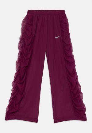 Fioletowe spodnie o szerokich nogawkach z marszczonymi tiulowymi falbanami po bokach i małym białym logo Nike Swoosh na przednim, prawym udzie.
