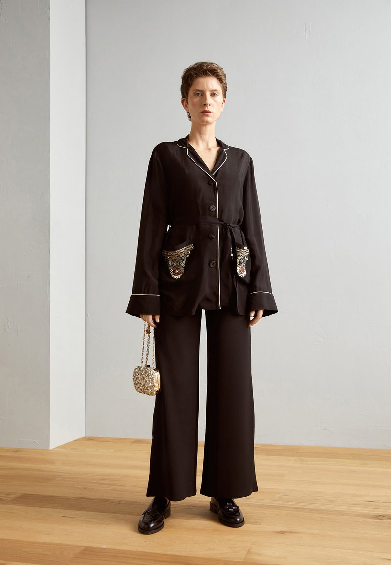 Vrouw die op een houten vloer staat, gekleed in een zwarte knoopjasje met geborduurde zakken, wijd broek, zwarte schoenen, en een kleine beaded handtas vasthoudt.