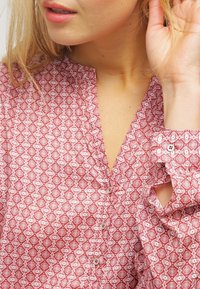 Blouse à motifs rouges avec un décolleté en V, fermeture à boutons et un design géométrique détaillé. Le tissu semble léger avec une texture lisse.