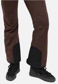 Brune tekniske bukser med sorte detaljer ved kanten, der har en glat overflade og en tætsiddende pasform. Kombinationen med sorte snørrede sneakers.