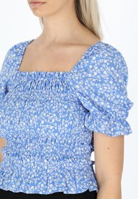 Blå blomstret bluse med firkantet halsudskæring, samlede stof, puffede ærmer og smock-design. Har hvidt blomstermønster og flæset kant.