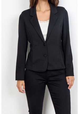Woman wearing a fitted black blazer with two buttons over a white lace-trimmed top and matching black pants against a plain background.