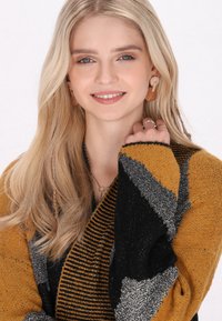 Maglione a maglia con motivo a zigzag nei colori nero, senape e argento. La modella indossa orecchini a cerchio e anelli, con lunghi capelli biondi.