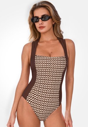 Vrouw in een bruin-beige badpak met geometrisch patroon en een zwarte zonnebril, poseert tegen een witte achtergrond.