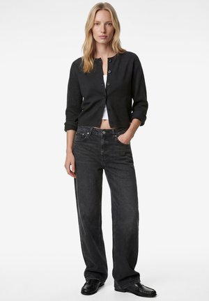 Vrouw draagt een zwarte cardigan met knopen over een witte cropped top, zwarte hoge taille wijd uitlopende jeans en zwarte loafers, staand met één hand in haar zak.