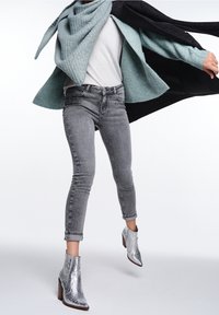 Jeans ajustados grises con acentos decorativos, un suéter gris claro y un cárdigan azul verde grande. Botas de tacón plateadas completan el look.