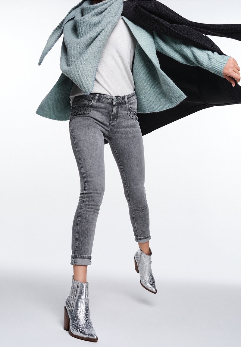 Jeans ajustados grises con acentos decorativos, un suéter gris claro y un cárdigan azul verde grande. Botas de tacón plateadas completan el look.