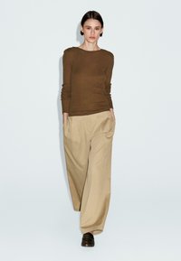 Brown fitted long-sleeve top with shoulder buttons paired with beige wide-leg trousers; model standing with hands in pockets.