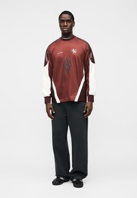 Haut de sport à manches longues bordeaux avec manches blanches et logo en forme de diamant, associé à un pantalon noir ample. Tissu texturé avec des détails de design sportif.