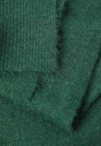 Dunkelgrüner Wollstoff mit weicher Textur, der feine Rippungen und lose Fasern aufweist, geschichtet mit einem gefalteten Kantendetail.