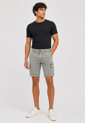 Man wearing black t-shirt, gray cargo shorts with rolled hems, and white sneakers, standing with one hand in pocket looking to side.