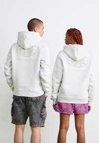Grå hættetrøjer med et logo på ryggen, der har kængurulommer. Den mandlige model bærer grå cargo shorts, mens den kvindelige model har lyserøde leopardmønstrede shorts på.
