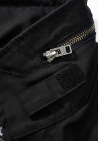 Černá tkanina oděvu s lesklým stříbrným kovovým zipem, suchým zipem a prošitými detaily. Makro snímek zobrazující texturu a detaily.