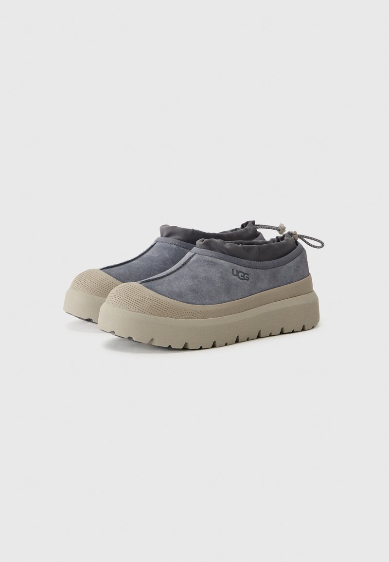 Chaussures slip-on en suède gris avec une semelle en caoutchouc beige plate et texturée. Présente une ouverture à cordon et un branding subtil sur le côté.