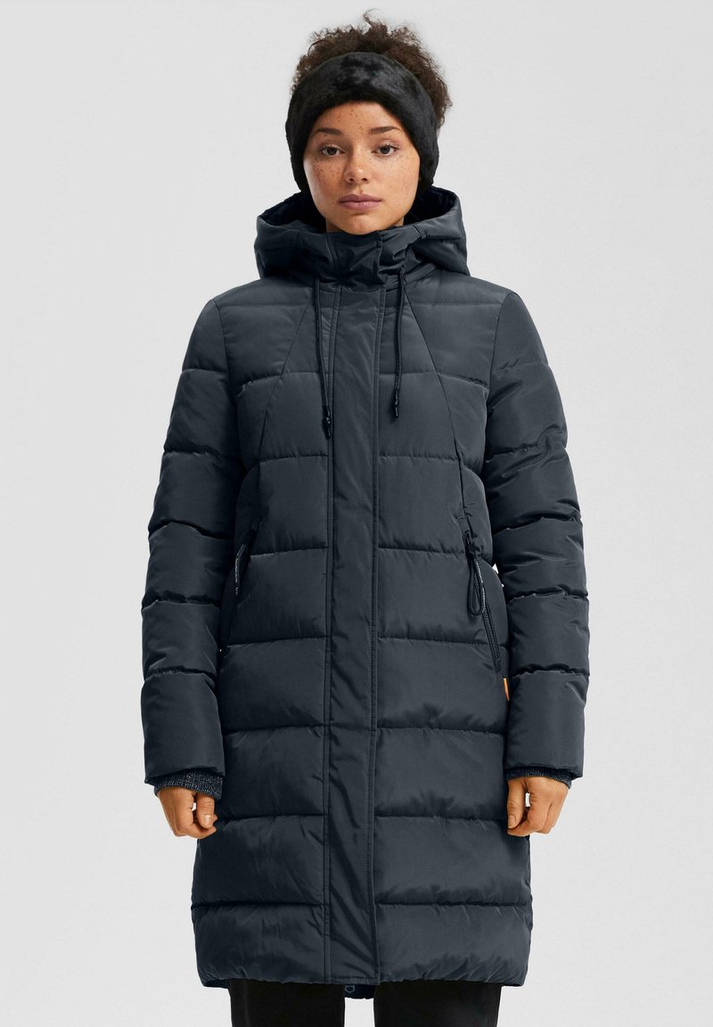 Moteris, vilkinti ilgą juodą šiltą žieminį paltą su gobtuvu ir juoda pūkuota galvos juosta, stovi prieš vienodą foną.