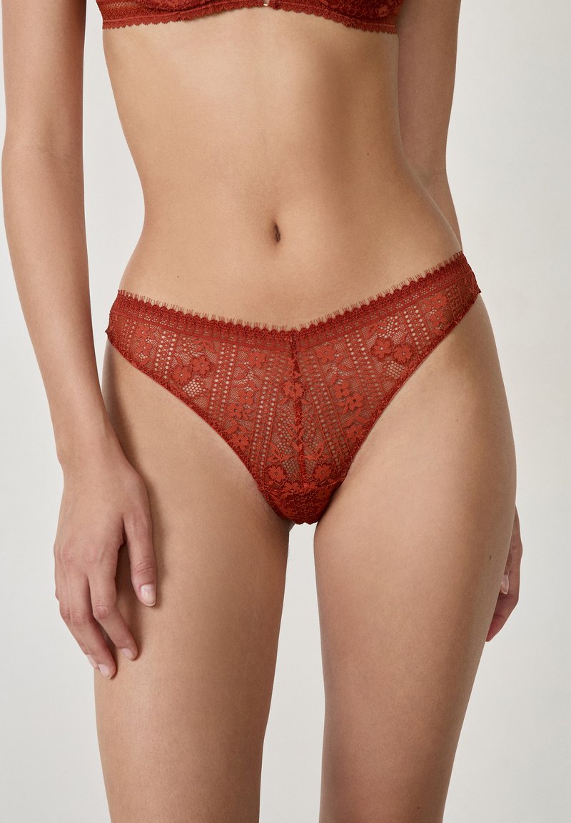 Lingerie en dentelle rouge avec motifs floraux, bords festonnés et texture semi-transparente, présentant une taille basse et des détails raffinés.