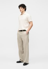 Άνδρας με ανοιχτό μπεζ πόλο, φαρδιά μπεζ παντελόνια με μαύρη ζώνη και μαύρα παπούτσια, όρθιος με τα χέρια στις τσέπες.