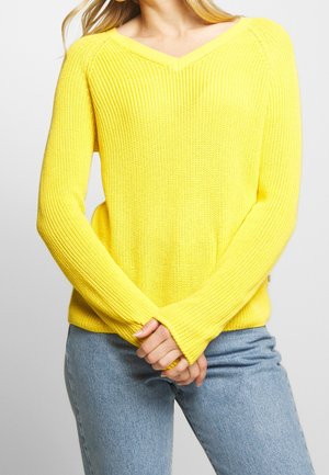 Kvinde iført en knaldrød gul strikket V-hals trøje med lange ærmer og lyseblå jeans, hænderne foldet foran.