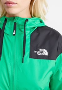 Vihreä ja musta tuulitakki, jossa on vetoketjukiinnitys, teksturoitu kangas, säädettävä huppu ja The North Face -logo rinnassa.