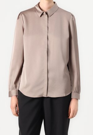 Donna che indossa una blusa taupe a maniche lunghe con bottoni nascosti e pantaloni neri su sfondo uniforme.