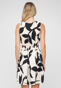 Mouwloze jurk in crème met zwart bladpatroon, voorzien van een smocktaille en een uitlopende rok, gemaakt van lichtgewicht stof.