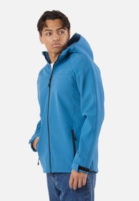 Giovane uomo che indossa una giacca blu con cappuccio e cerniera, con le mani in tasca, che guarda di lato, su uno sfondo bianco semplice.