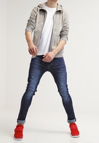 Giacca grigia con cappuccio sopra una t-shirt bianca, jeans blu e calzature rosse. L'outfit presenta una vestibilità rilassata e colori a contrasto.