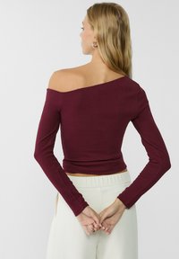 Burgunderfarbener, schulterfreier Langarm-Top mit tailliertem Design, dehnbarem Material und cropped Länge, kombiniert mit hellen Hosen.
