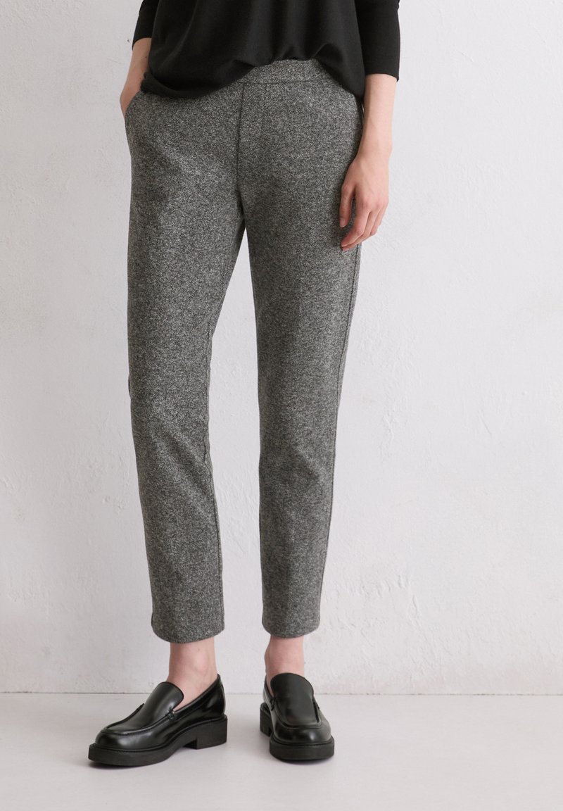Graue Wollmischungshosen mit einem schmal zulaufenden Design und Seitentaschen, kombiniert mit schwarzen Leder-Slippern, vor einem neutralen Hintergrund.