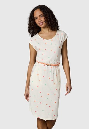 Kvinne med krøllete hår iført en hvit knelengde kjole med flerfargede hjerte-mønstre og et tynt oransje belte.