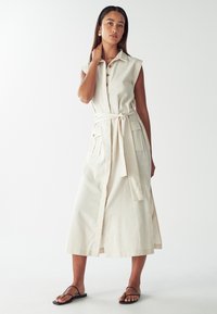 Donna che indossa un abito midi beige senza maniche, con cintura e tasche, e sandali neri, in piedi contro uno sfondo bianco.