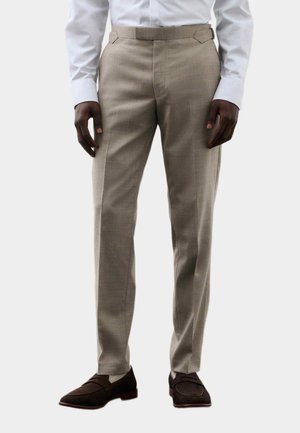Mann trägt beige Stoffhose, weißes langärmeliges Hemd und dunkelbraune Slipper, steht vor einem einfarbigen Hintergrund.