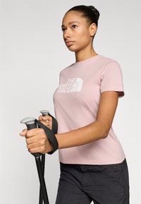 Ανοιχτό ροζ μπλουζάκι με το λογότυπο "The North Face", μαζί με μαύρα μπαστούνια πεζοπορίας με γκρι λαβές. Το μοντέλο φοράει μαύρο σορτς.