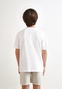 Camiseta blanca de algodón con mangas cortas y escote redondo, con una pequeña etiqueta rectangular en la parte central de la espalda. Llevada con pantalones cortos beige.