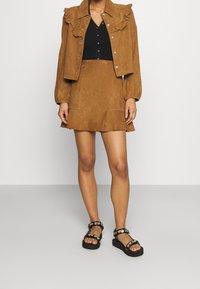 Brown suede jacket with ruffled collar, paired with a matching skirt. Black top and chunky black sandals with metal accents complete the outfit.