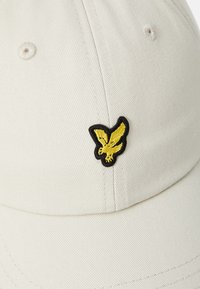 Beige baseball-lippis, jossa on teksturoitu kangas. Edessä musta-keltainen brodeerattu lintulogo. Näkyvissä kuusi tuuletusaukkoa.