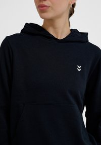 Vrouw die een zwarte hoodie draagt met een klein wit dubbel-chevron logo op de borst, waarbij een deel van haar gezicht en zilveren oorringen zichtbaar zijn.