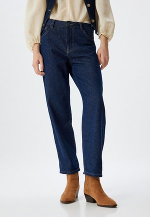 Vaqueros de denim azul con un diseño de pierna recta, que presentan un estilo clásico de cinco bolsillos y costuras amarillas en contraste. Se llevan con botas marrones hasta el tobillo.