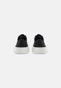 Schwarze Canvas-Schuhe mit niedrigem Design, weißen Gummisohlen und strukturierten Akzenten. Die Rückansicht zeigt gepolsterte Kragen und Nähdetails.