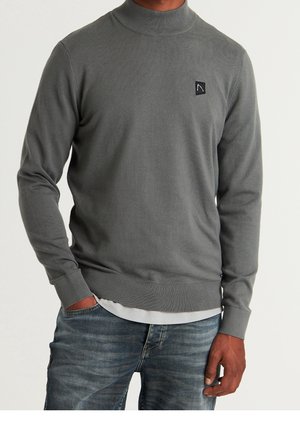 Pull en tricot gris avec un col montant, des poignets et un ourlet côtelés, comportant un petit patch logo noir sur la poitrine. Tissu doux et léger.