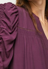 Blusa viola con maniche a sbuffo in tessuto arricciato, delicati dettagli in pizzo sul collo e una vestibilità morbida.