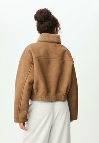 Hnědá umělá kožená zkrácená bunda s měkkým, huňatým límcem, elastickým lemem a prošitím na ramenech. Nositá přes šedé kalhoty.