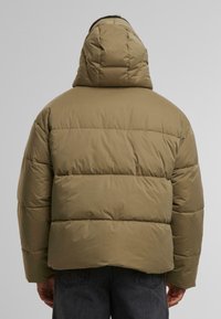 Pufferjakke i olivengrønn med hette, med en kviltet design og kontrasterende sydd linjer; laget av et glatt, lett stoff.