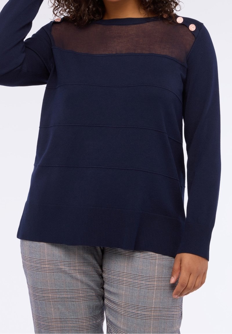 Femme portant un pull marine avec un empiècement transparent et trois boutons dorés sur l'épaule, associé à un pantalon gris à carreaux.