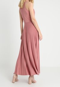 Długa, różowa sukienka bez rękawów z dopasowanym gorsetem i plisowanym dołem. Wykonana z gładkiego materiału, sięga do podłogi. Do sukienki dołączone są sandały na obcasie z paskami.