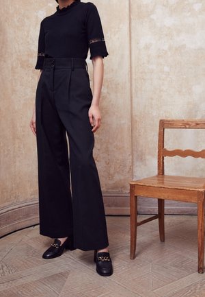 Top nero a maglia con rifiniture in pizzo, abbinato a pantaloni neri a vita alta e gamba larga e mocassini neri con tacco a blocco e dettaglio in catena dorata.