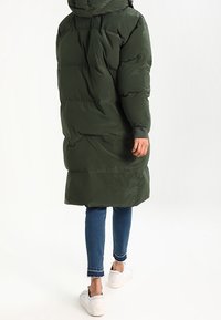 Długa, puchowa ciemnozielona kurtka z kapturem, z poziomymi przeszyciami. Połączona z dopasowanymi niebieskimi dżinsami i białymi trampkami.
