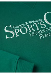 Tissu vert avec texte brodé blanc "Healthy & Wellness SPORTS LES REVOIR France" et bord-côte visible.